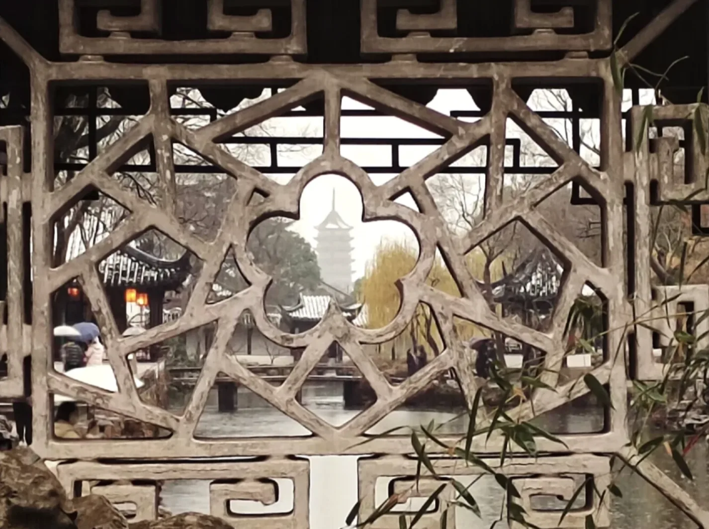 镂空石窗框住雨中古镇河道与远处宝塔 / A carved stone window framing a rainy old-town canal and distant pagoda