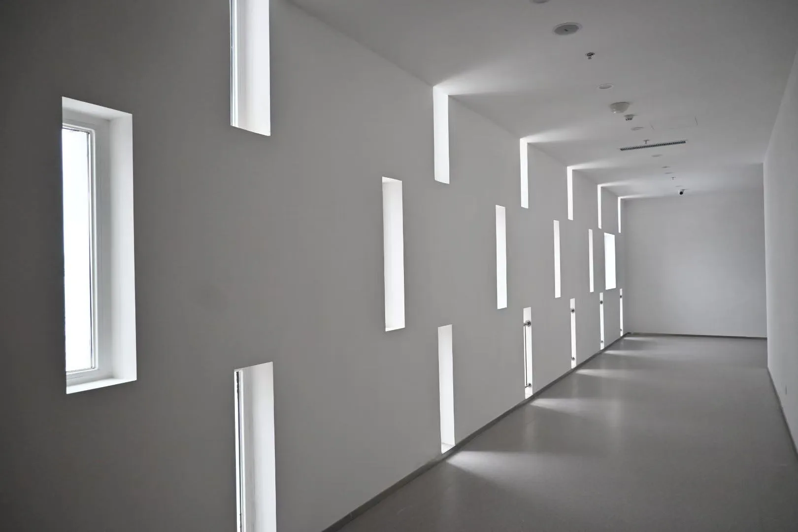 白色走廊一侧排列着高低错落的狭长窗户 / A white corridor with staggered narrow windows